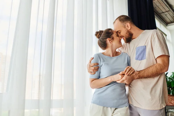 Vista lateral de una pareja sonriente en ropa de casa cogida de la mano y abrazándose mientras está de pie en casa - foto de stock