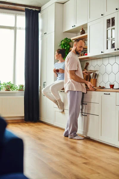 Man in homewear taking plates with tasty breakfast near girlfriend holding tea in kitchen at home — Stock Photo