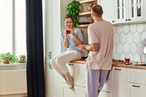 Улыбающаяся женщина в домашней одежде держит чай рядом с парнем с завтраком на тарелке на кухне утром — стоковое фото