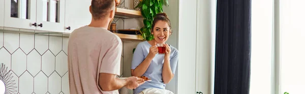 Sorridente donna bruna in loungewear che tiene il tè vicino al fidanzato con colazione in cucina, banner — Foto stock