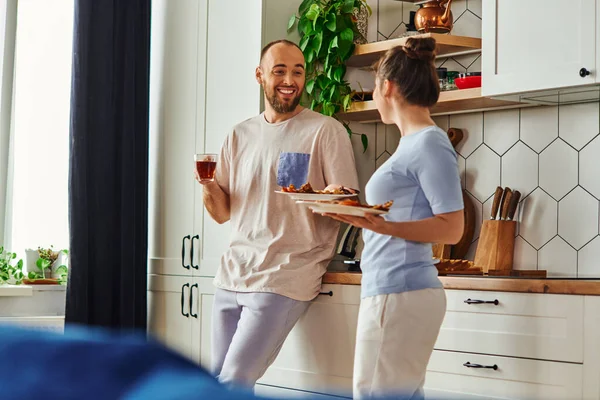 Homem alegre em loungewear segurando chá e conversando com a namorada com café da manhã em pratos em casa — Fotografia de Stock