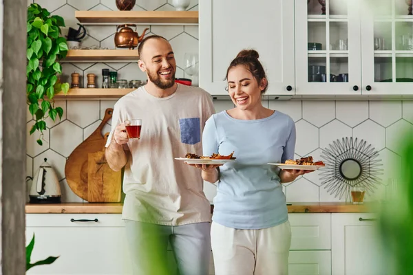 Femme souriante dans les vêtements de maison tenant des assiettes avec petit déjeuner près du petit ami avec thé dans la cuisine — Photo de stock