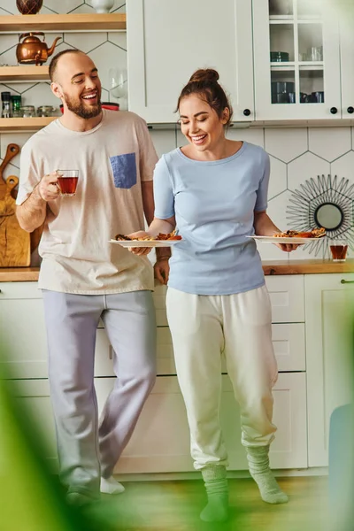 Smiling couple in homewear holding tasty breakfast on plates and tea in morning in kitchen at home — Stock Photo