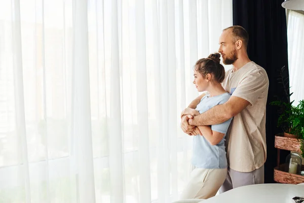 Side view of bearded man in homewear hugging girlfriend while standing near window at home — Stock Photo