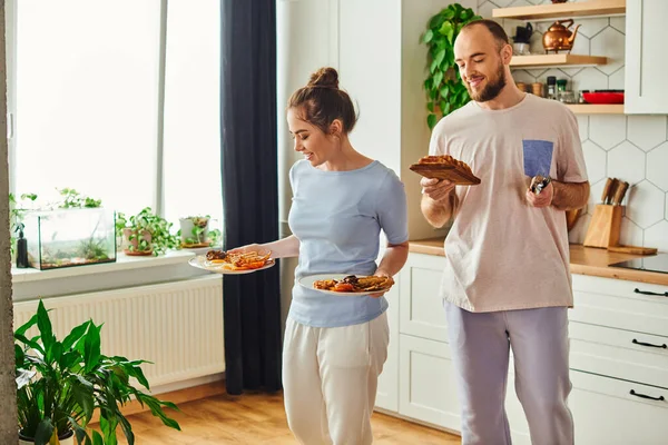 Улыбающаяся пара в домашней одежде держит тосты и тарелки с вкусным завтраком дома утром — стоковое фото