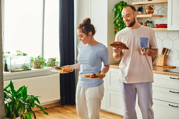 Coppia sorridente in biancheria da casa che tiene brindisi e piatti con gustosa colazione a casa al mattino — Foto stock