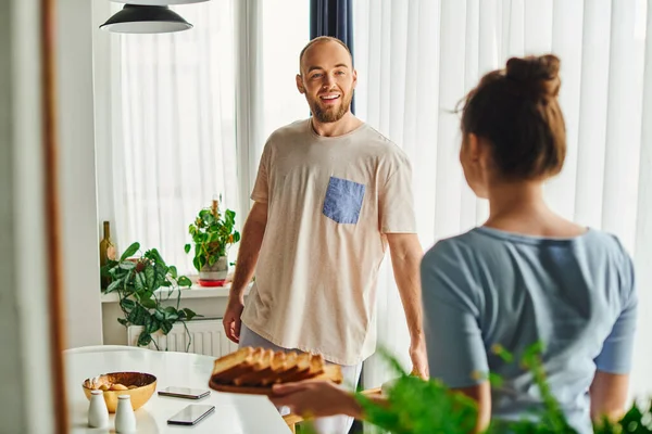 Sorrindo homem em roupa de casa de pé perto namorada borrada com café da manhã caseiro em casa — Fotografia de Stock
