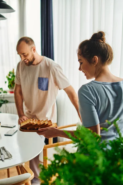 Smiling woman in homewear holding toasts near blurred boyfriend during breakfast at home in morning — Stock Photo