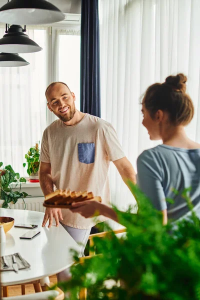 Smiling man standing near smartphones and blurred girlfriend with breakfast at home in morning — Stock Photo