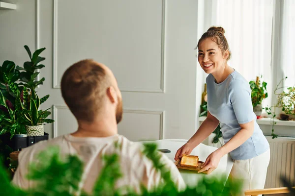 Fröhliche Frau in Hauskleidung stellt Toasts auf den Tisch neben verschwommenem Freund beim Frühstück zu Hause — Stockfoto