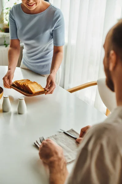 Улыбающаяся женщина кладет тосты на стол рядом с размытым парнем и столовыми приборами во время завтрака дома — стоковое фото