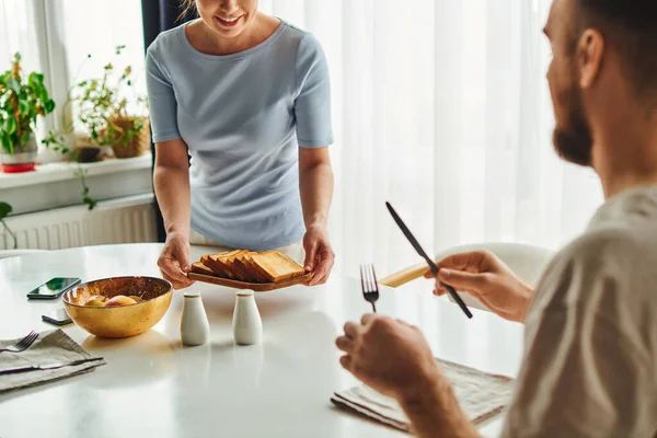Улыбающаяся женщина кладет тосты на стол рядом с бойфрендом со столовыми приборами и смартфонами утром дома — стоковое фото