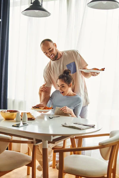 Sorrindo homem colocando café da manhã na mesa perto de namorada animada e smartphone em casa de manhã — Fotografia de Stock