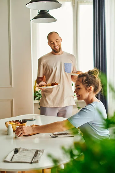 Donna sorridente che prende il sale vicino al fidanzato offuscato con la colazione sui piatti a casa al mattino — Foto stock
