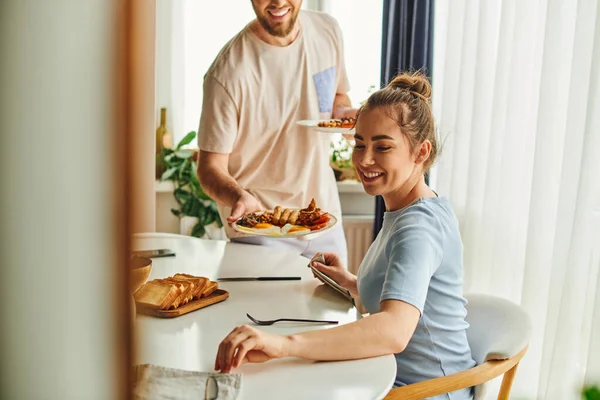 Sorrindo mulher sentada perto de talheres e namorado borrado segurando saboroso café da manhã em casa de manhã — Fotografia de Stock