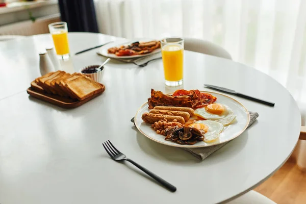 Pequeno-almoço saboroso com feijão e ovos fritos na mesa perto de talheres e suco de laranja em casa — Fotografia de Stock