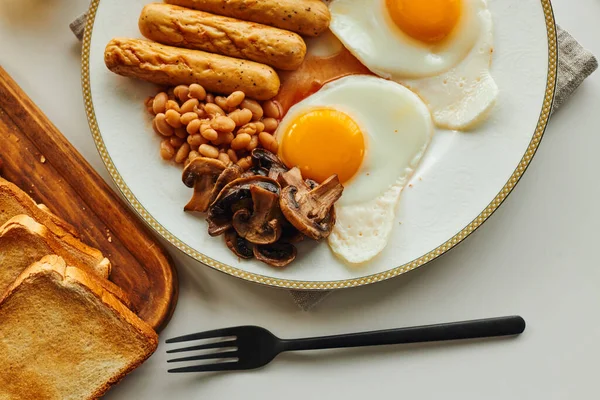 Top view of breakfast with fried eggs and sausages near toasts on cutting board on table at home — Stock Photo