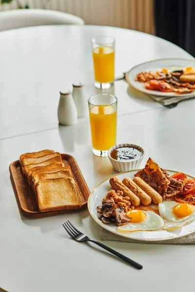 Colazione tradizionale con uova fritte, salsicce e pancetta vicino al succo d'arancia e marmellata sul tavolo a casa — Foto stock