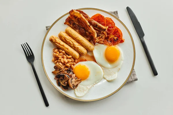 Вид зверху ранкового прийому їжі з сосисками, смаженими яйцями та беконом на тарілці біля столових приборів на столі — стокове фото