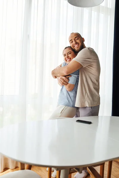 Усміхнений бородатий чоловік в домашньому одязі обіймає дівчину біля смартфона на столі вдома — стокове фото