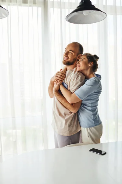 Mujer sonriente en ropa de casa abrazando novio y de pie cerca de teléfono inteligente con pantalla en blanco en casa - foto de stock