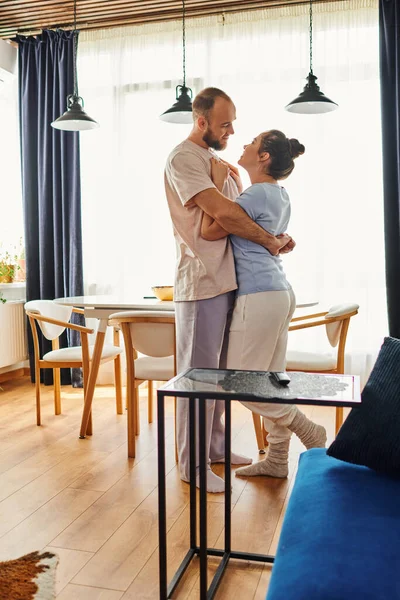Hombre barbudo en ropa de casa abrazando a su novia sonriente mientras está de pie en la sala de estar en casa - foto de stock