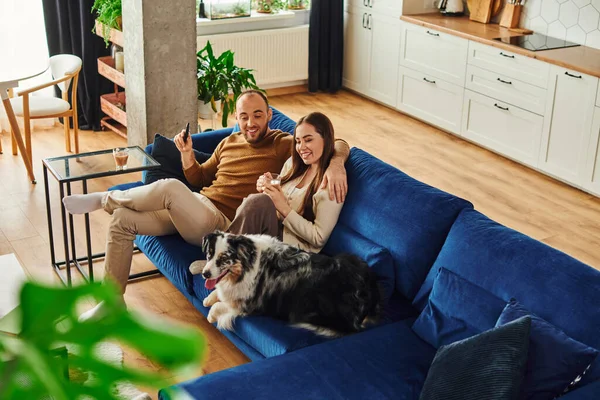 Усміхнена пара з віддаленим контролером і кавою, що сидить біля кордону колі на дивані вдома — стокове фото