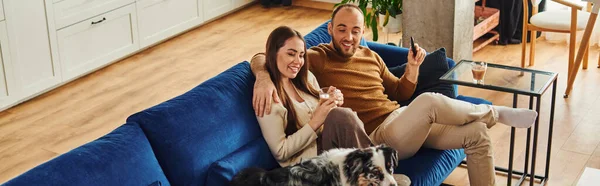 Coppia sorridente con caffè e telecomando seduta vicino al bordo collie sul divano a casa, banner — Foto stock