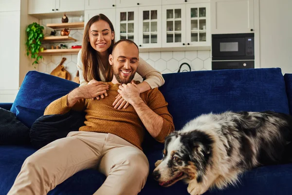 Joyful woman hugging boyfriend in casual clothes and looking at border collie on couch at home — Stock Photo