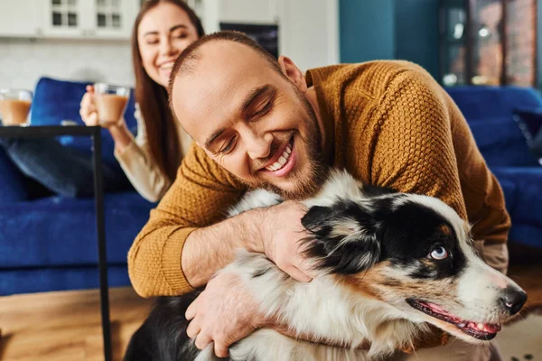 Homme joyeux étreignant frontière collie chien près de petite amie floue avec café dans le salon à la maison — Photo de stock
