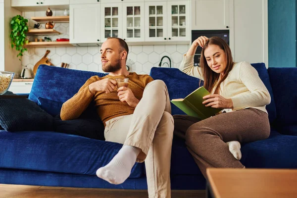 Man holding cup of coffee while smiling girlfriend reading book and spending time on couch at home — Stock Photo