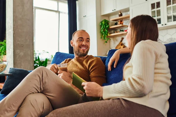 Sonriente hombre sosteniendo una taza de café y hablando con su novia con libro mientras está sentado en el sofá en casa - foto de stock