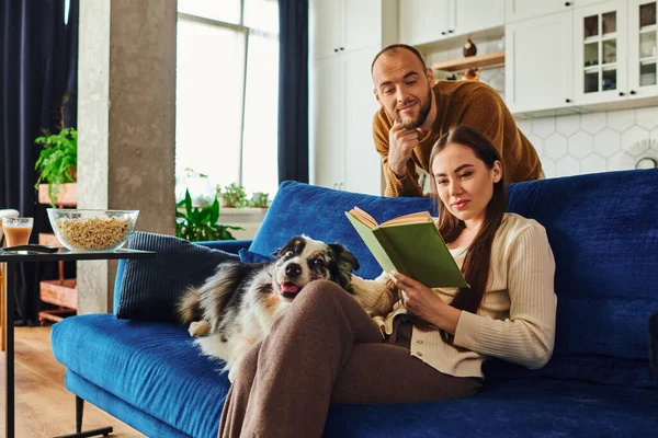 Livre de lecture femme près du petit ami et frontière collie chien sur vouvh dans le salon à la maison — Photo de stock