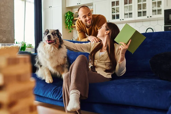 Усміхнена пара проводить час з книгою і прикордонною собакою на дивані біля попкорну у вітальні — стокове фото