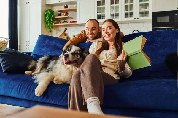 Casal sorridente com livro relaxante perto de fronteira collie no sofá e pipoca na sala de estar em casa — Fotografia de Stock