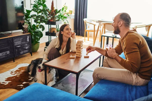 Smiling man holding coffee and playing wood blocks game with girlfriend near border collie at home — Stock Photo