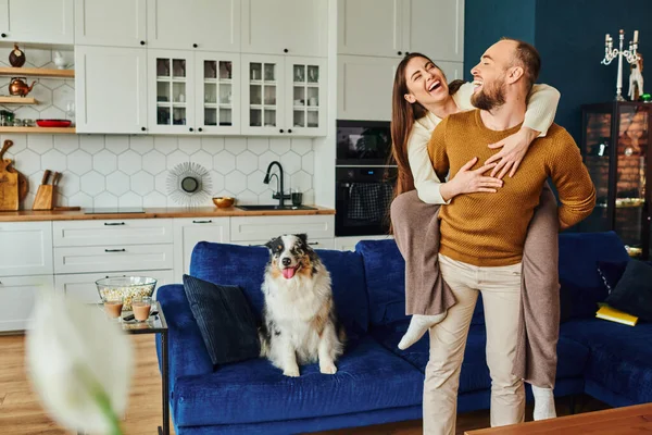 Smiling woman piggybacking on boyfriend near border collie dog on couch and popcorn in living room — Stock Photo