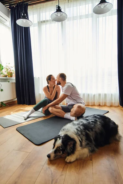 Smiling couple in sportswear kissing and sitting on fitness mats near blurred border collie at home — Stock Photo