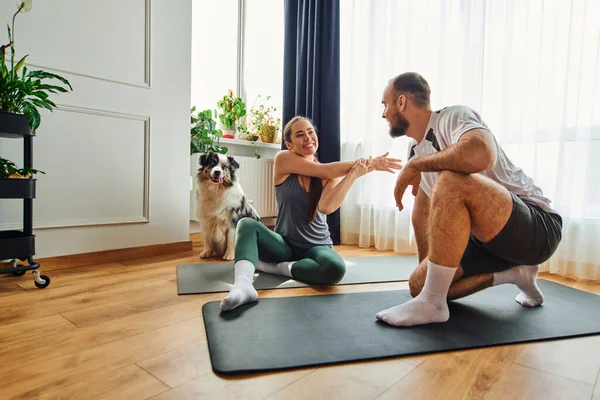 Mulher sorridente em sportswear aquecendo no tapete de fitness perto de namorado e border collie em casa — Fotografia de Stock