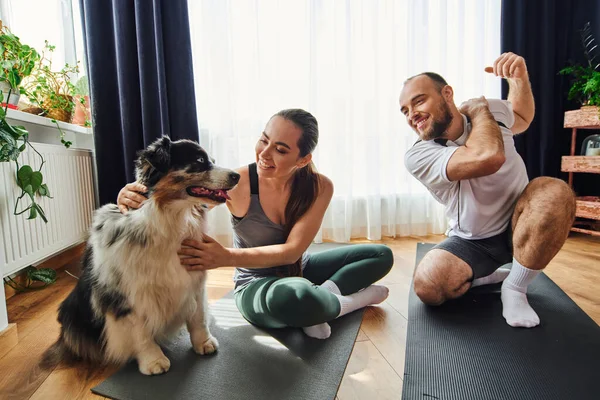 Smiling man warming up near girlfriend in sportswear petting border collie on fitness mat at home — Stock Photo