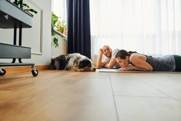Uomo sorridente in abbigliamento sportivo guardando fidanzata e confine collie mentre sdraiato sul tappeto fitness a casa — Foto stock