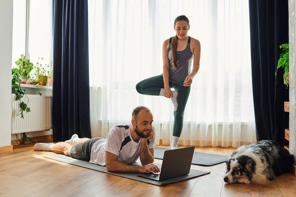 Sorrindo mulher de pé na esteira de fitness, enquanto namorado usando laptop perto de border collie em casa — Fotografia de Stock