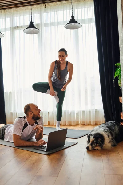 Donna positiva in piedi sul tappeto fitness vicino al fidanzato utilizzando laptop e border collie a casa — Foto stock