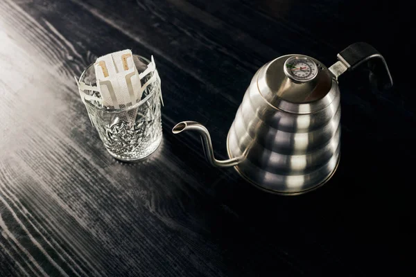 Método pour-over, hervidor de agua de goteo metálico, vidrio con café molido en bolsa de filtro sobre mesa negra - foto de stock
