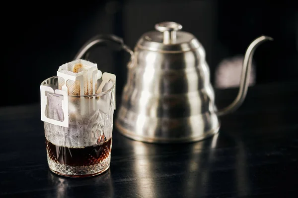Glass with paper filter bag and black expresso near blurred drip kettle on black table, drip method — Stock Photo