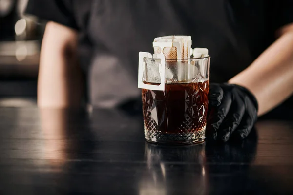 Обрізаний вид на бариста пивоваріння ароматичної кави в паперовому мішку фільтра і кришталеве скло на чорному столі — стокове фото