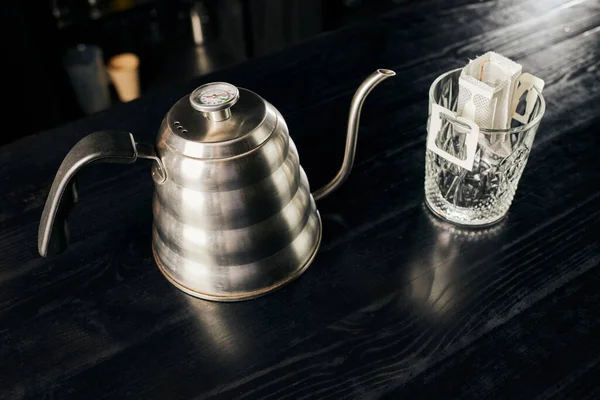 Metallic drip kettle, crystal glass with ground coffee in filter bag on black table, drip method — Stock Photo