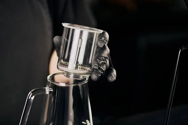 Coffee shop, partial view of barista in black latex glove holding siphon coffee maker above glass pot — Stock Photo