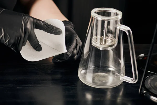 Обрізаний вид на бариста в чорних латексних рукавичках, що тримає паперовий фільтр біля кавоварки з сифоном — стокове фото