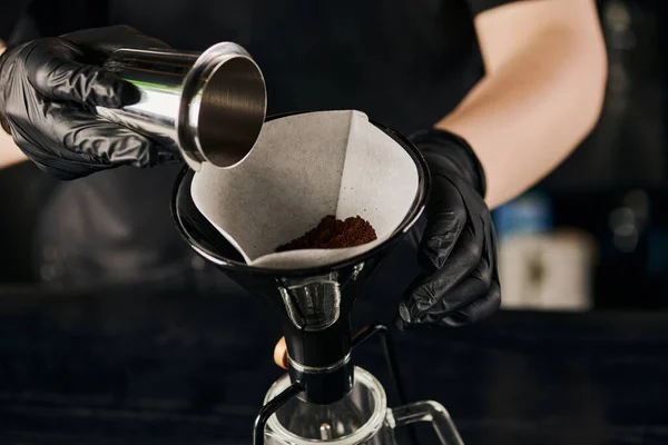 Barista in lattice nero guanti versando caffè macinato da jigger nel filtro della caffettiera sifone — Foto stock
